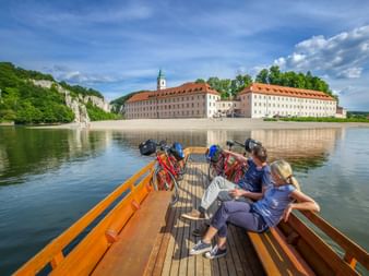 Radfahrer auf Holzfähre überqueren die Donau in Richtung Kloster Weltenburg mit seinen markanten weißen Gebäuden und orangen Dächern.