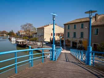 Blue pedestrian bridge crossing Canal du Midi in Homps, with traditional French houses and a moored boat along the waterfront under clear blue sky.