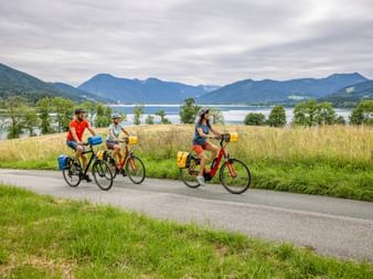 Three cyclists with yellow panniers riding on paved path beside Lake Tegernsee, with mountains and cloudy sky in background.