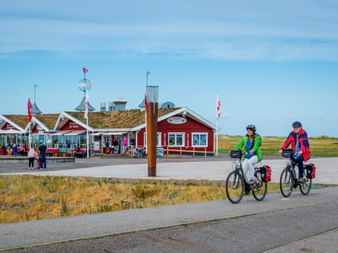 Zwei Radfahrer mit Tourenrädern und Packtaschen fahren an roten Holzstrandgebäuden in St. Peter-Ording unter blauem Himmel vorbei.