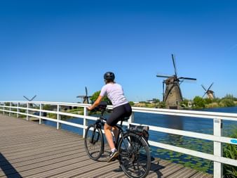 Female cyclist on wooden bridge with white railings at Kinderdijk, viewing traditional Dutch windmills across the canal under clear blue sky.