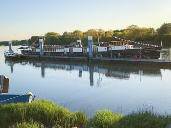 Flusskreuzfahrtschiff MS Magnifique III liegt bei Sonnenuntergang auf ruhigem Wasser, geschmückt mit Flaggen. Grünes Flussufer im Vordergrund.