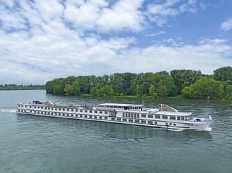 White MS Rigoletto river cruise ship sailing on a wide waterway in North Holland and Friesland, with green tree-lined shores under blue sky.