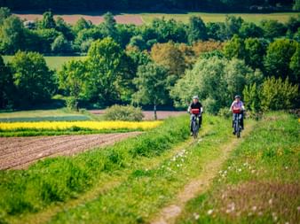 Two cyclists riding on a rural path through green fields in Weserbergland. Rolling hills with trees and farmland visible in the background.