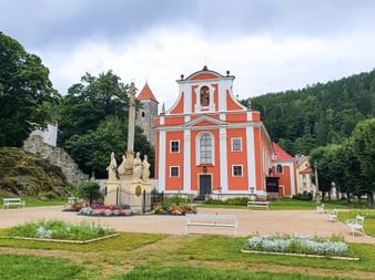 Orange baroque church Maria Heimsuchung with white trim in Bohemia, surrounded by green lawns, flower beds, and forested hills.