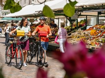 Two cyclists with e-bikes walking through a farmers market in Bolzano with colorful fruit and vegetable stands under white canopies.