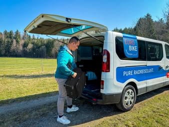 Mitarbeiter in türkisfarbener Jacke lädt Gepäck in weißen Eurobike-Transporter mit offener Hecktür in ländlicher Umgebung mit grünen Feldern und Wald.