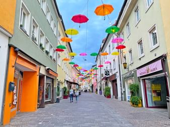 Fußgängerzone in Villacher Altstadt mit bunten Schirmen über der Straße. Mehrfarbige Gebäude mit Geschäften säumen beide Seiten.
