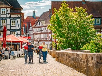 Zwei Radfahrer schieben ihre Räder über den Kopfsteinplatz in Melsungen, umgeben von traditionellen Fachwerkhäusern und Straßencafé.