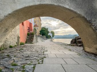 View through a stone arch in Passau showing cobblestone path leading to the Inn River, with colorful historic buildings and the Schaiblingsturm tower visible.