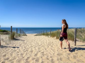 Female cyclist in sportswear holding helmet, standing on sandy dune path leading to beach with wooden fence posts and dune grass under blue sky.