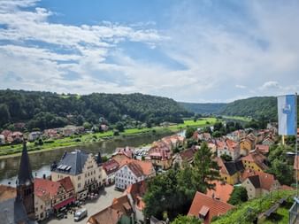 Luftaufnahme von Stadt Wehlen mit historischen Gebäuden und roten Ziegeldächern entlang der Elbe, umgeben von grünen Hügeln und Wäldern.