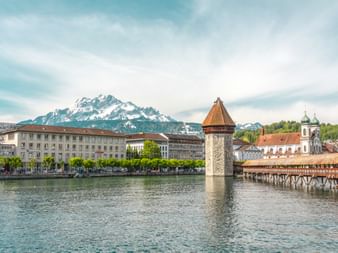 Kapellbrücke mit Wasserturm an der Reuss in Luzern, mit historischen Gebäuden am Ufer und schneebedeckten Bergen im Hintergrund.
