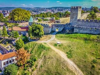 Luftaufnahme der Festung Kalemegdan in Belgrad mit Steinmauern, Türmen und grünen Hängen. Stadtskyline im Hintergrund unter blauem Himmel.