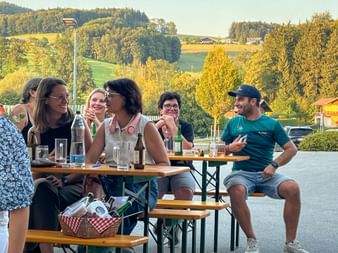 Gruppe von Personen sitzt an Holz-Biergartentischen im Freien, genießt Getränke und Gespräche an einem Sommerabend mit ländlicher Landschaft im Hintergrund.