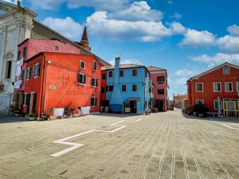 Gepflasterter Platz in Pellestrina, umgeben von bunten Häusern in Orange, Blau, Rosa und Rot. Ein Kirchturm erhebt sich hinter den Gebäuden unter blauem Himmel.