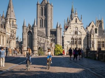 Zwei Radfahrer auf Kopfsteinpflasterplatz in Gent mit St.-Nikolaus-Kirche und historischen Gebäuden. Fußgänger unter blauem Himmel.