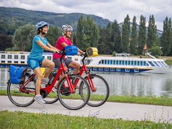 Two cyclists on the riverbank with a ship in the background