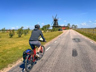 Cyclist with panniers riding on paved road toward traditional windmill in rural Swedish landscape under blue sky.