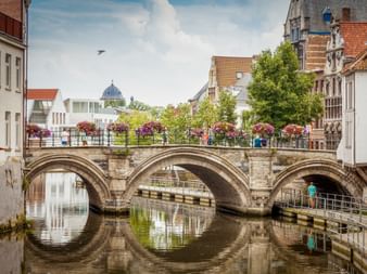Historische Steinbrücke Grootbrug mit drei Bögen in Mechelen, geschmückt mit bunten Blumenkörben, spiegelt sich im ruhigen Wasser.