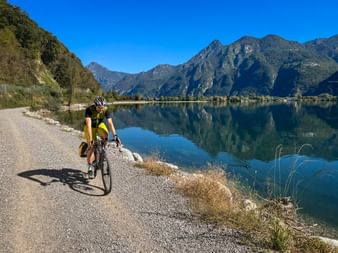 Radfahrer im gelben Trikot auf Schotterweg am Idrosee mit Bergen, die sich im ruhigen Wasser unter blauem Himmel spiegeln.