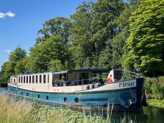 MS Fleur, a blue and white river cruise ship, moored along a tree-lined canal. The vessel features rows of cabin windows and an open deck area.