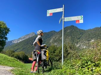 Radfahrer mit Tourenrad und Packtaschen an Wegweiser mit Routen nach Salzburg und Weißbach, mit bewaldeten Bergen unter blauem Himmel.