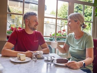 Two cyclists in sports shirts enjoying cake and coffee at Café Zauner in Bad Ischl. Large windows show flower boxes and greenery outside.