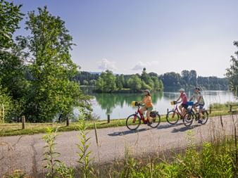 Three cyclists riding along a paved path beside a calm green river with trees and forested hills in the background under blue sky.