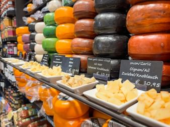 Auslage bunter Käselaibe in Reihen gestapelt mit Kostproben darunter. Preisschilder zeigen Sorten wie Zwiebel/Knoblauch für €10,95 in Gouda.