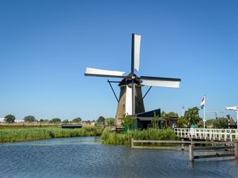 Traditionelle holländische Windmühle mit weißen Flügeln neben einem Kanal in Kinderdijk. Eine weiße Holzbrücke überquert das Wasser mit Schilf und niederländischer Flagge.