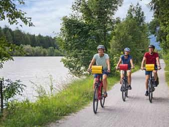 Three cyclists with helmets and colorful panniers riding along the Ruhr Valley Cycle Route beside a river with trees and green landscape.