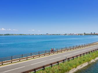 Two cyclists on a coastal bridge with blue lagoon waters and Grado cityscape in the distance under clear blue sky.