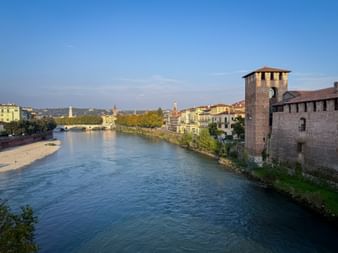 View of the Adige River in Verona with Castelvecchio's medieval tower on the right and Ponte Scaligero bridge in the background under clear sky.