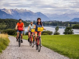 Three cyclists with helmets riding on a gravel path near Riegsee lake. Alpine mountains and green meadows visible in the background under cloudy sky.