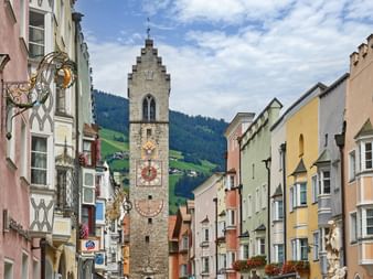 Stone church tower with ornate clock in Sterzing, framed by colorful buildings. Green mountain slopes visible in background under cloudy sky.