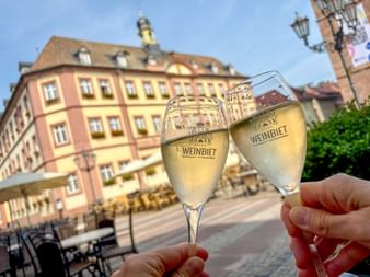 Zwei Hände stoßen mit Weingläsern mit der Aufschrift 'Weinbiet' vor historischen Gebäuden am Neustadter Marktplatz an.