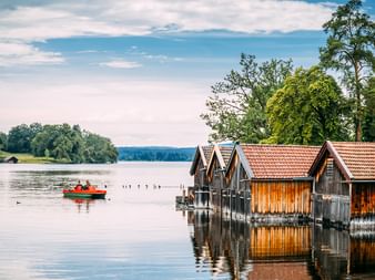 Wooden boathouses with red tile roofs on Staffelsee lake. A red pedal boat floats nearby. Trees line the shore under a blue sky.