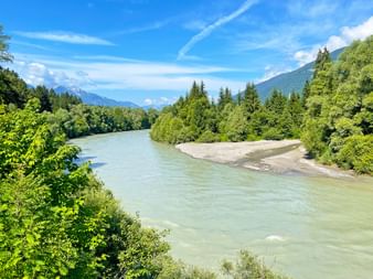 Türkisfarbene Drau fließt durch grüne Waldlandschaft bei Steinfeld. Berge im Hintergrund unter blauem Himmel mit weißen Wolken.