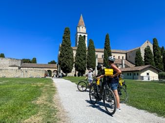 Two cyclists with yellow panniers on a gravel path leading to Aquileia Basilica with its tall bell tower and cypress trees under blue sky.