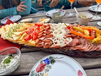 Wooden platter with Austrian cold cuts, cheese, vegetables and bread on rustic table. People sharing meal with floral plates and drinks.