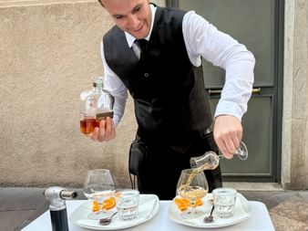 Waiter in black vest pouring digestif into glasses on white plates with orange garnish and chocolates at an outdoor table in Trient.
