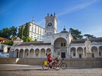 Zwei Radfahrer fahren über einen Kopfsteinpflasterplatz in Udine, vorbei an weißem Arkadengebäude mit Glockenturm und Statue unter blauem Himmel.