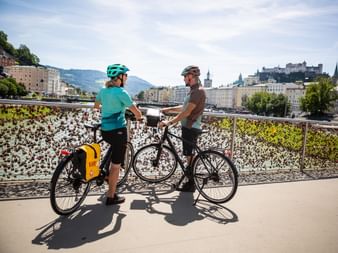 Two cyclists with helmets standing on a pedestrian bridge in Salzburg, with Hohensalzburg Fortress and historic buildings in background.
