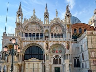 Prunkvolle Fassade des Markusdoms in Venedig mit gotischen Bögen, Türmen, Kuppeln und dekorativen Skulpturen unter blauem Himmel.