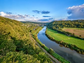 Luftaufnahme der Weser, die sich durch grünes Tal bei Trendelburg schlängelt. Bewaldete Hügel, Wiesen und ein Dorf unter blauem Himmel.