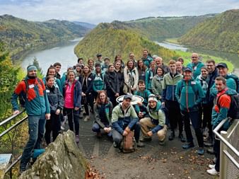 Large group of Eurohike team members in hiking gear posing at a scenic viewpoint overlooking the Danube River with forested hills and mountains.