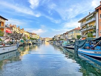 Kanal im Hafen von Grado, gesäumt von bunten Gebäuden und Fischerbooten, die an beiden Seiten unter blauem Himmel mit Schleierwolken liegen.