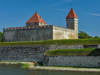 Cyclist riding past Kuressaare Castle with stone walls and red-tiled towers. The medieval fortress sits on a green hill under blue sky.