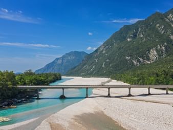 Bridge spanning the turquoise Tagliamento River near Venzone with white gravel banks and green mountains in the background under blue sky.